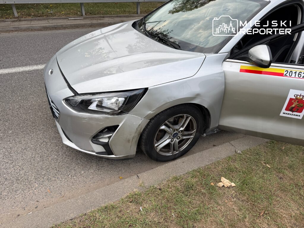 Uszkodzony samochód osobowy zaparkowany na poboczu drogi, z wgniecioną maską i otwartymi drzwiami.