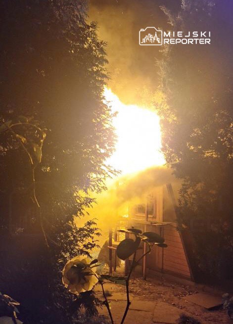 Ogień wydobywający się z budynku otoczonego drzewami, emitujący gęsty dym w ciemnej scenerii nocnej.