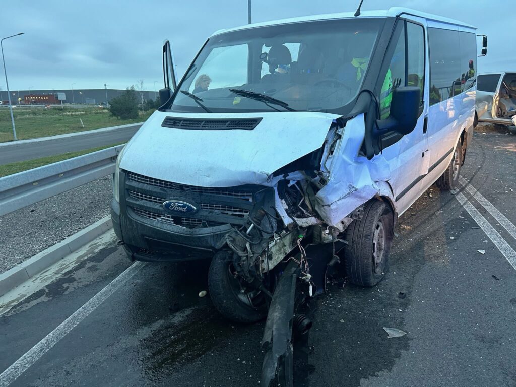 Rozbity furgon na drodze, z uszkodzoną przednią częścią i otwartymi drzwiami, obok widać inne pojazdy.