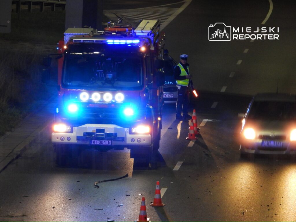 Straż pożarna z niebieskimi światłami zatrzymuje ruch na drodze, a policjant kieruje samochodami obok miejsca wypadku.