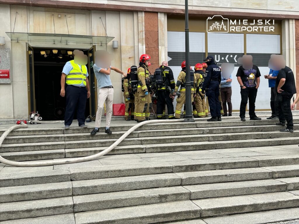 Grupa strażaków w hełmach i mundurach rozmawia z policjantami przed wejściem do budynku, obok leży wąż strażacki.