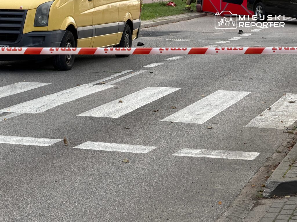 Żółty samochód dostawczy stoi na przejściu dla pieszych, otoczone taśmą ostrzegawczą. Na drodze leżą liście.