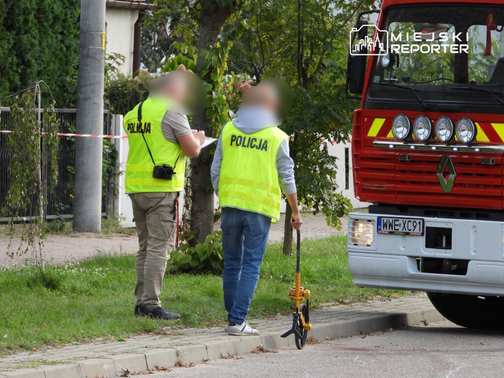 Policjanci w kamizelkach odblaskowych rozmawiają przy czerwonym wozie strażackim, badając miejsce zdarzenia.