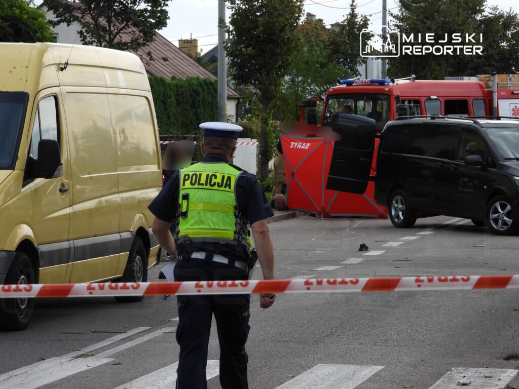 Policjant w kamizelce odblaskowej kieruje ruchem na zablokowanej drodze, obok czerwony wóz strażacki i żółty furgon.