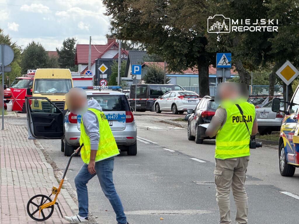 Policjant w odblaskowej kamizelce mierzy odległość na ulicy, obok parkowane samochody i pojazdy służb ratunkowych.