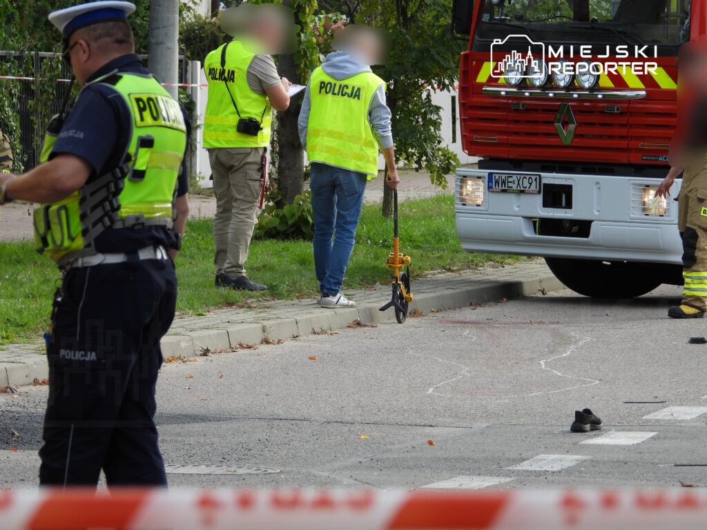 Policjanci zabezpieczają miejsce zdarzenia, na którym znajduje się ślad krwi i porzucone buty, obok stoi wóz strażacki.