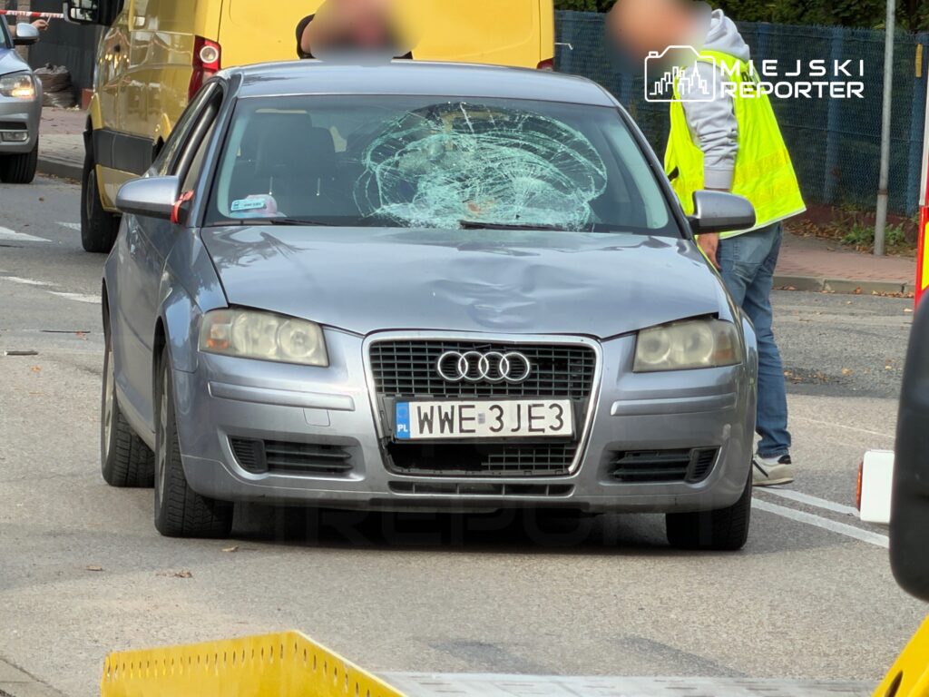 Srebrne audi z rozbitą przednią szybą stoi na ulicy, obok pracownicy w kamizelkach odblaskowych prowadzą działania.