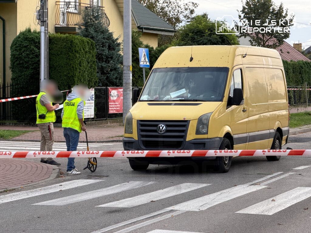 Policjanci w kamizelkach odblaskowych prowadzą pomiary w miejscu zdarzenia, obok żółtego furgonu, otoczeni taśmą zabezpieczającą.