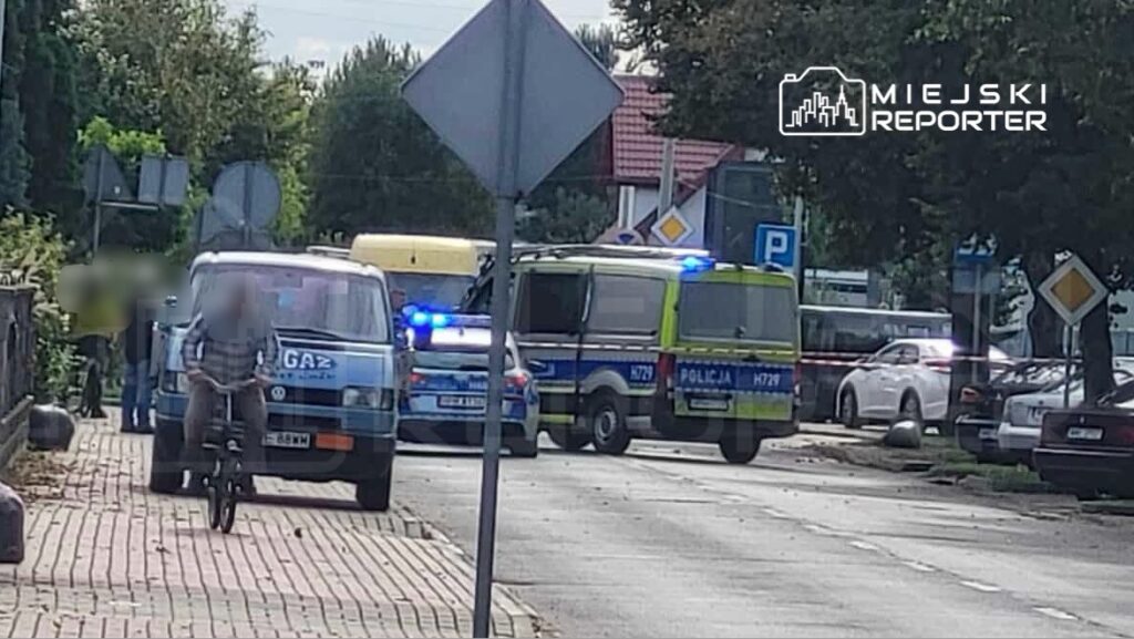 Mężczyzna na rowerze przejeżdża obok policyjnych radiowozów i furgonetek z włączonymi sygnałami świetlnymi.