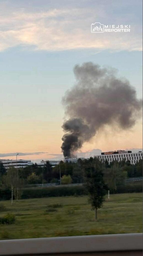 Czarny dym unoszący się w powietrzu nad nowoczesnym biurowcem w późnym popołudniu.