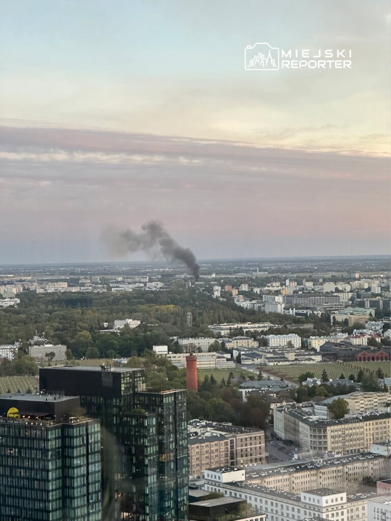 Czarny dym unoszący się nad zielonym terenem w mieście, w tle widoczne nowoczesne budynki i czerwony komin.