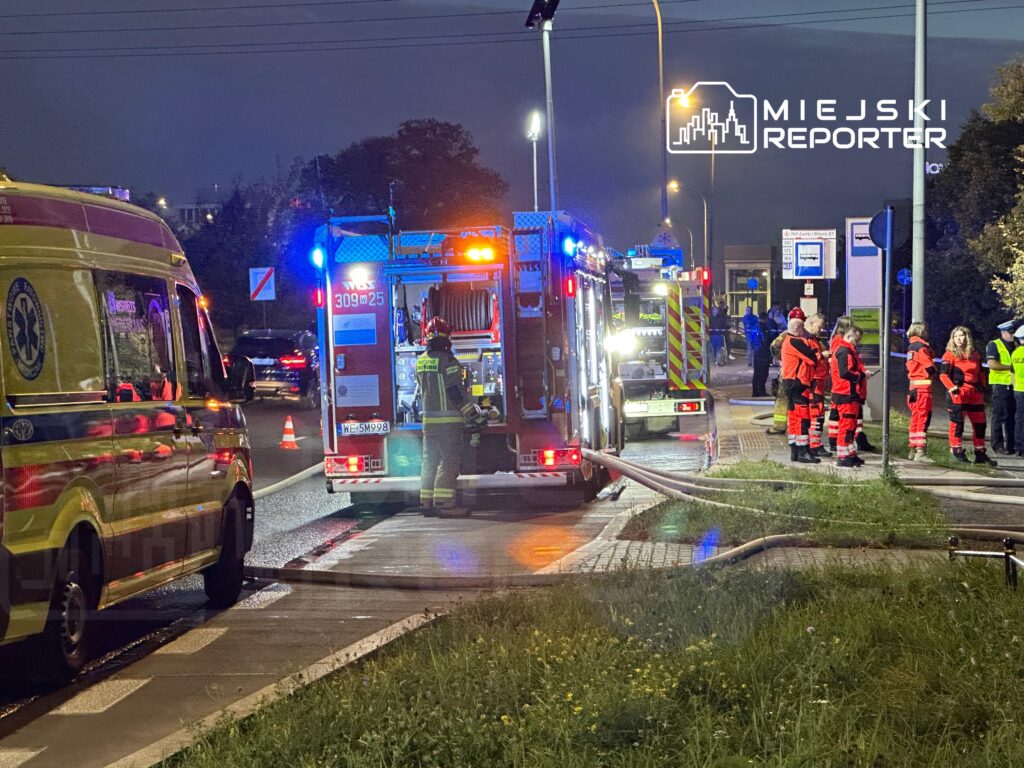 Ratownicy w mundurach omawiają sytuację obok wozu strażackiego na oświetlonej ulicy.