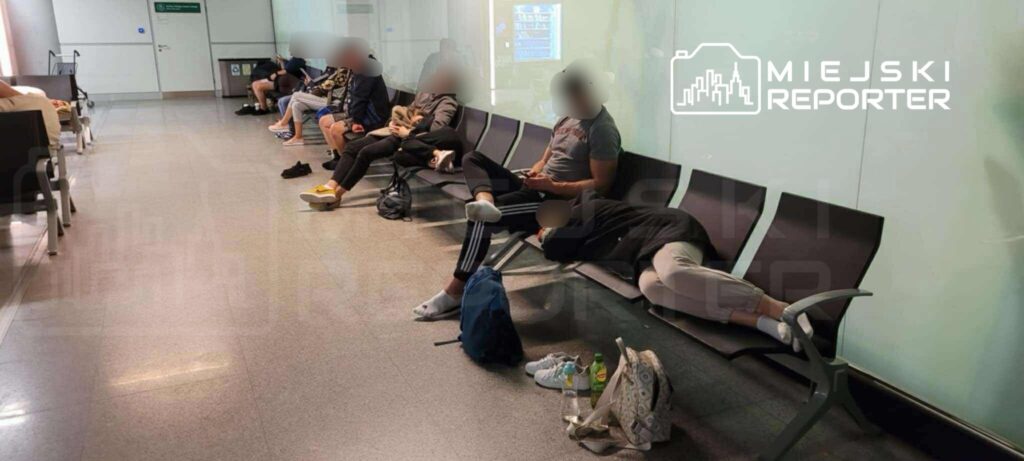 Grupa osób siedzi na ławkach w poczekalni, niektórzy korzystają z telefonów, a inni odpoczywają.