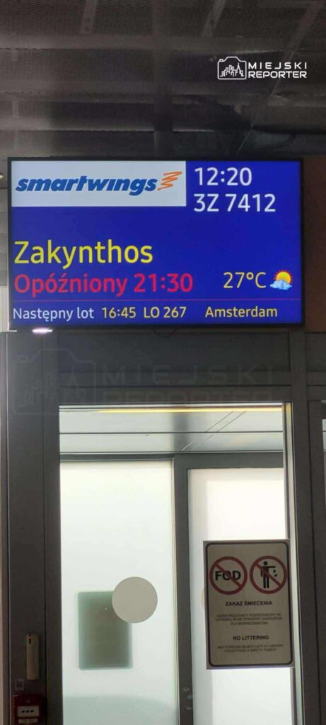 Tablica informacyjna na lotnisku, wskazująca opóźnienie lotu do Zakynthos oraz szczegóły kolejnego lotu do Amsterdamu.