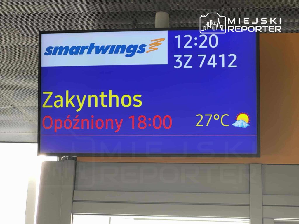 Tablica informacyjna na lotnisku z danymi o locie do Zakynthos, opóźnionym o godzinę, z temperaturą 27°C.