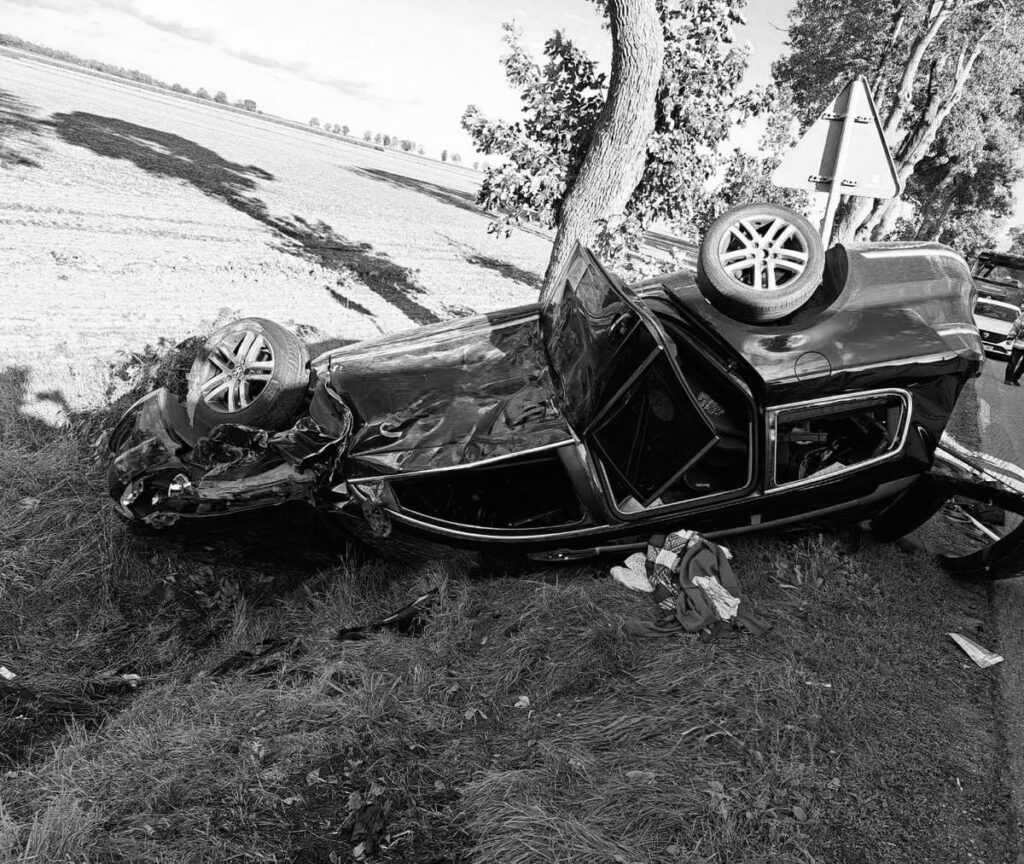Zniszczony samochód leży na dachu obok drogi, otoczony trawą i drzewami, z widoczną uszkodzoną karoserią.