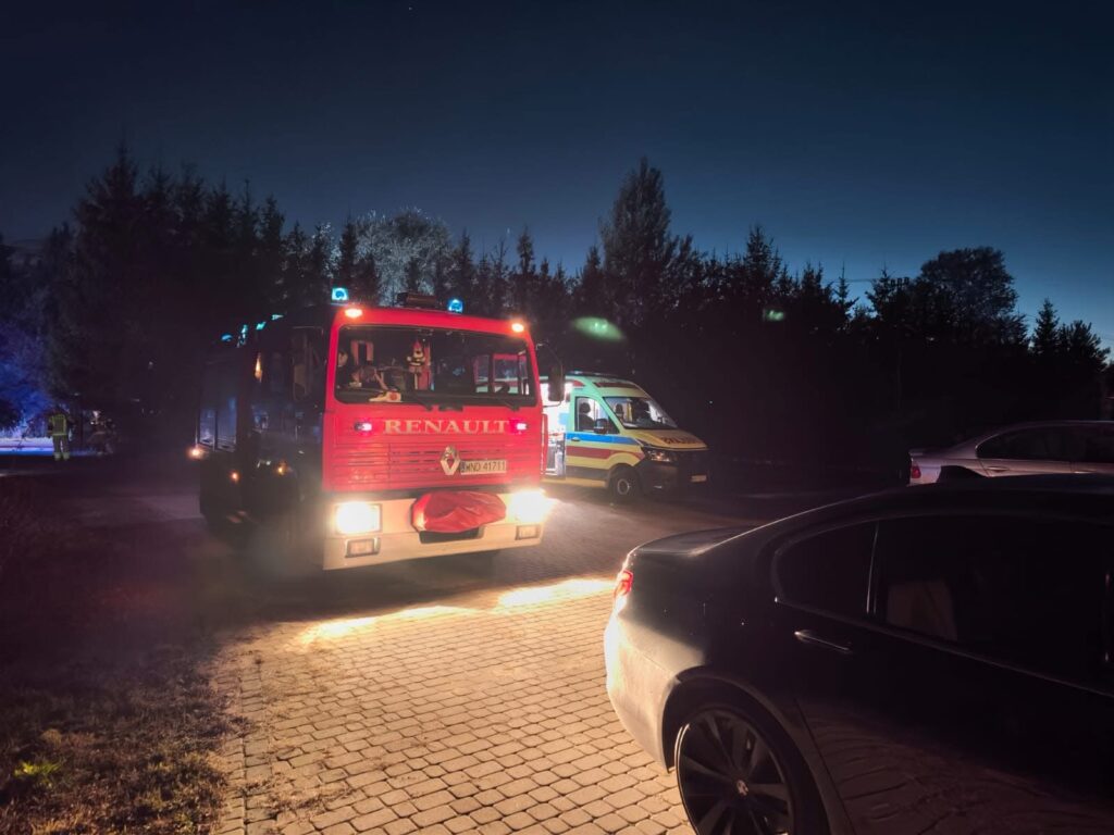 Czerwony wóz strażacki z niebieskimi światłami parkowany na drodze, obok karetka pogotowia w nocy.