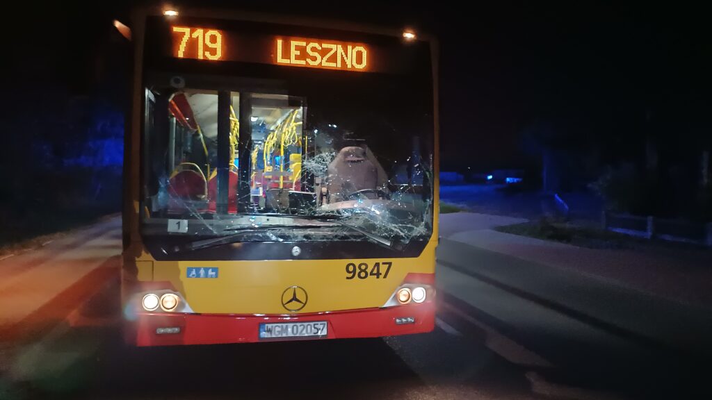 Autobus linii 719 z rozbitą szybą stoi na ulicy nocą, oświetlony przez latarnie i niebieskie światła.