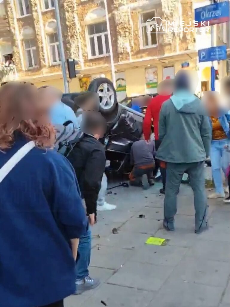 Grupa osób otacza przewrócony samochód osobowy na ulicy, niektórzy próbują pomóc.