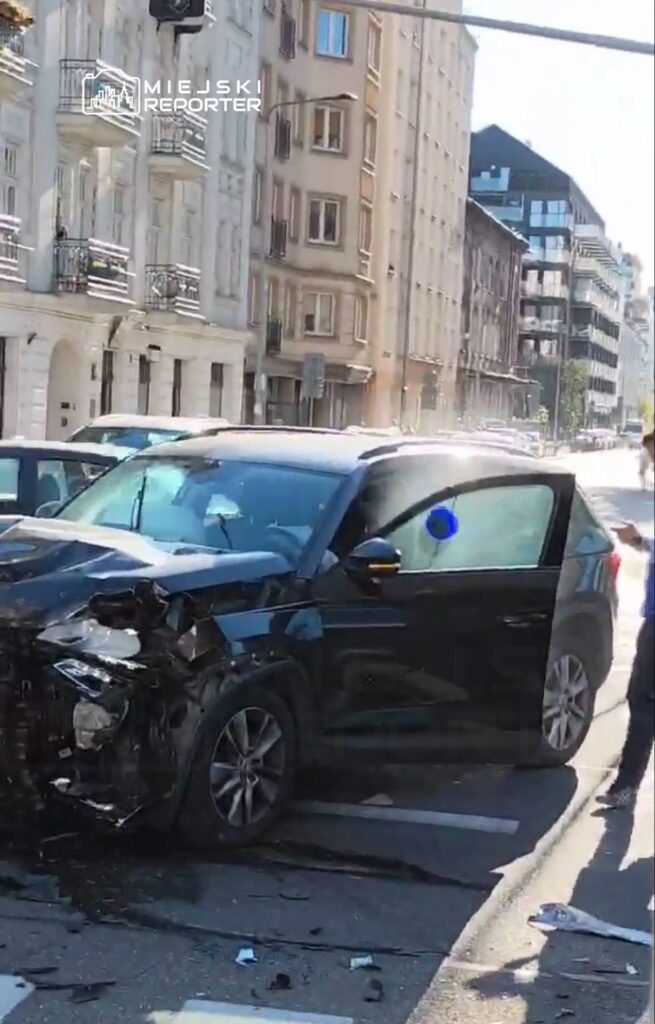 Czarny samochód osobowy z uszkodzoną przednią częścią stoi na ulicy, obok dwóch innych pojazdów.