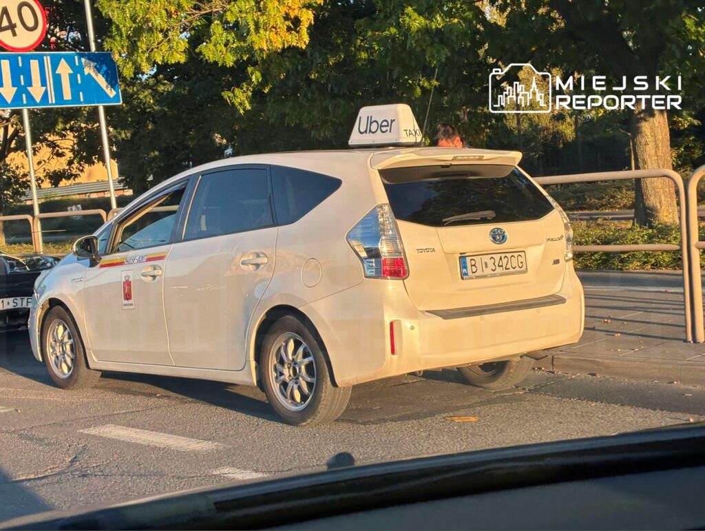 Biały samochód osobowy z logo Ubera zatrzymany na ulicy przy znaku ograniczenia prędkości do 40 km/h.