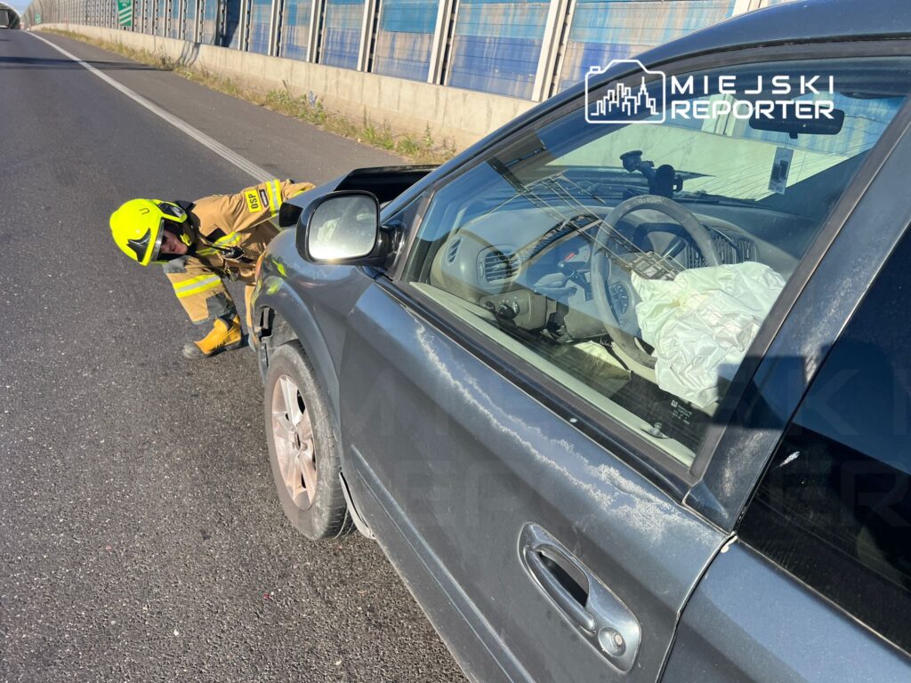 Strażak w hełmie bada uszkodzony samochód na poboczu drogi, obok widoczne zniszczenia pojazdu.