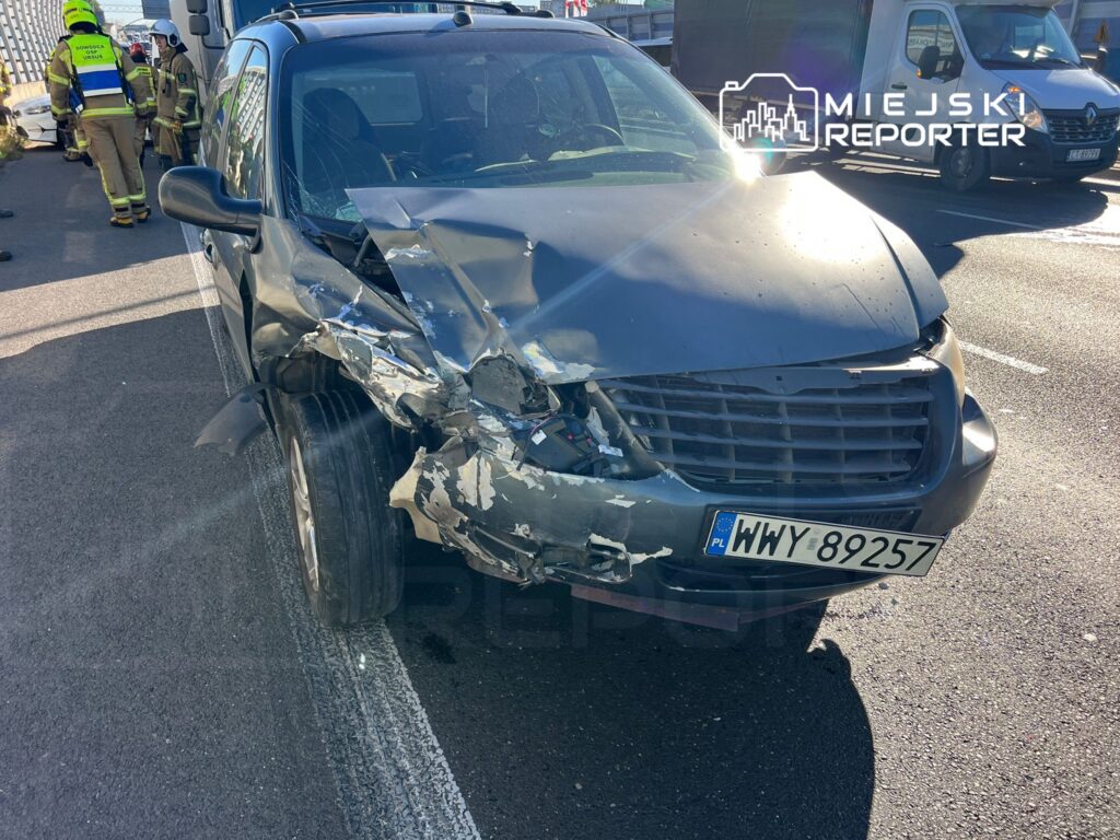Zniszczony samochód osobowy z uszkodzoną przednią częścią stoi na drodze, obok grupy ratowników.