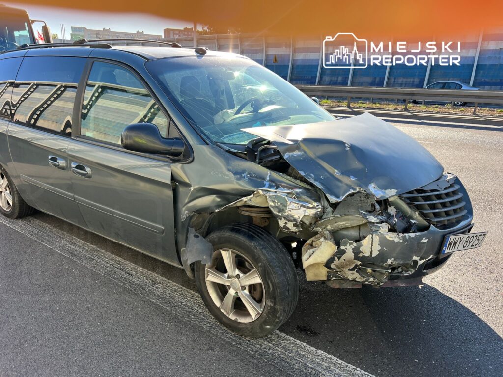 Uszkodzony minivan z wgniecionym przodem stoi na pasie ruchu, obok widoczne inne pojazdy na drodze.