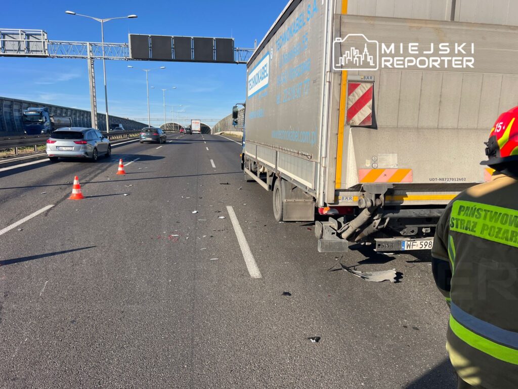 Na drodze leżą resztki zderzenia, a w tle stoi ciężarówka, obok której pracują strażacy w mundurach.