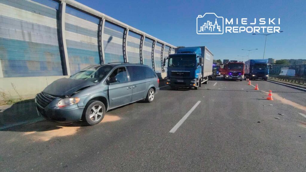 Samochód osobowy z uszkodzoną maską stoi na drodze, obok ciężarówki i pojazdów służb ratunkowych, na ruchliwej jezdni.