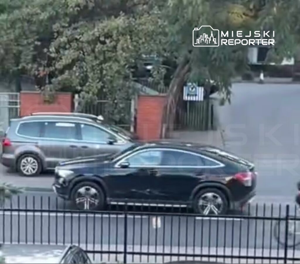 Czarny SUV przejeżdża obok zaparkowanego szarego samochodu na ulicy otoczonej zielenią.