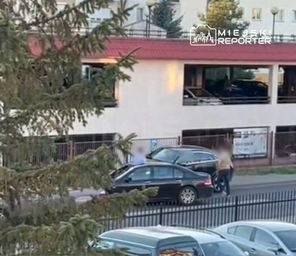 Dwie osoby rozmawiają obok zaparkowanych samochodów na ulicy, w tle widoczny budynek z przeszkloną fasadą.