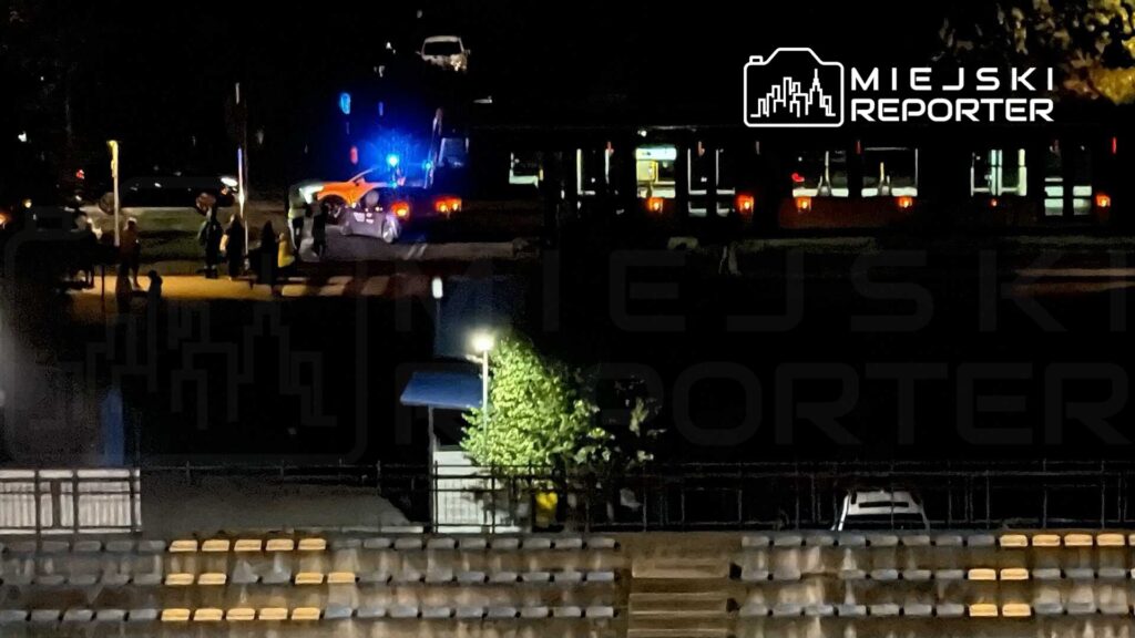 Grupa ludzi czeka na przystanku, podczas gdy policyjny radiowóz z włączonymi sygnałami świetlnymi stoi obok autobusu.