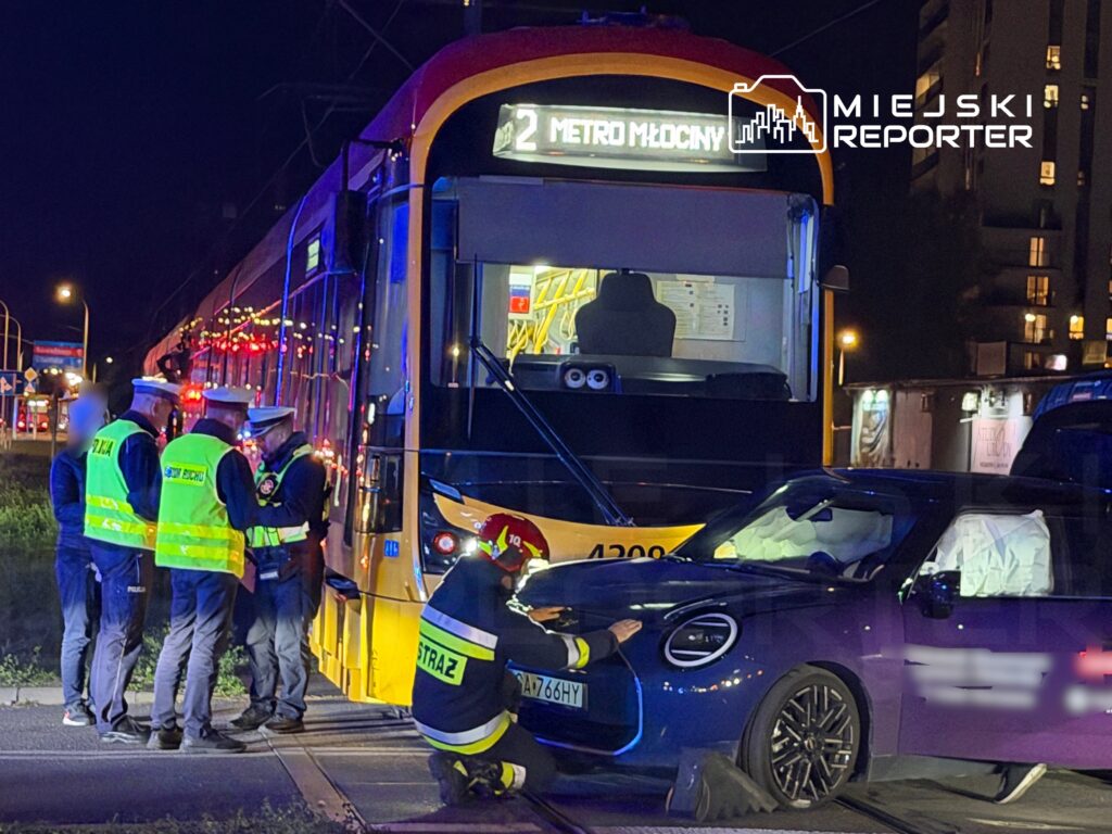 Grupa policjantów i strażaków bada uszkodzony samochód osobowy przy torach tramwajowych w nocy, obok tramwaju.