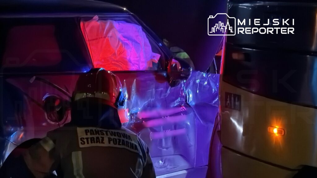 Strażak w hełmie bada uszkodzenia samochodu osobowego o zgniecionej karoserii w nocy, otoczony przez niebieskie światła.