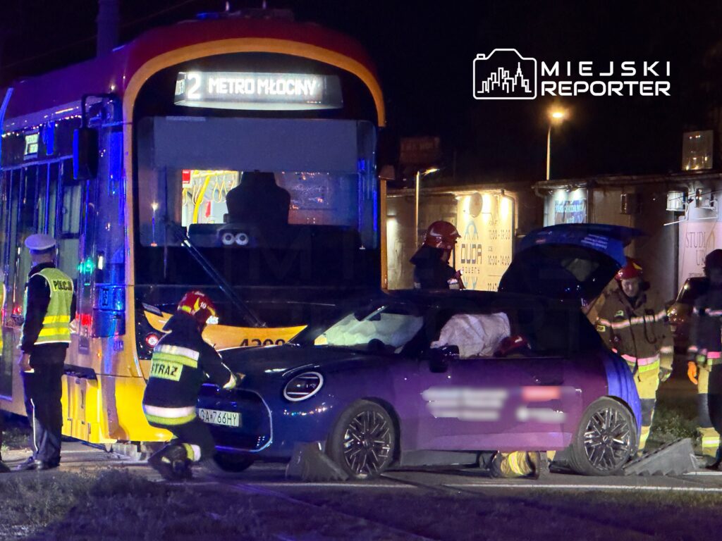 Kierowca samochodu osobowego leży w pojeździe, a strażacy oraz policja pracują przy wypadku drogowym z udziałem tramwaju.