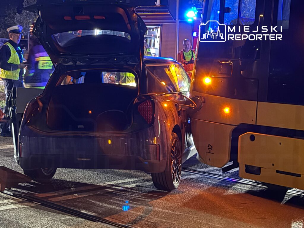 Czarny samochód osobowy z otwartym bagażnikiem stoi blisko tramwaju, a obok pracują policjanci i strażacy.