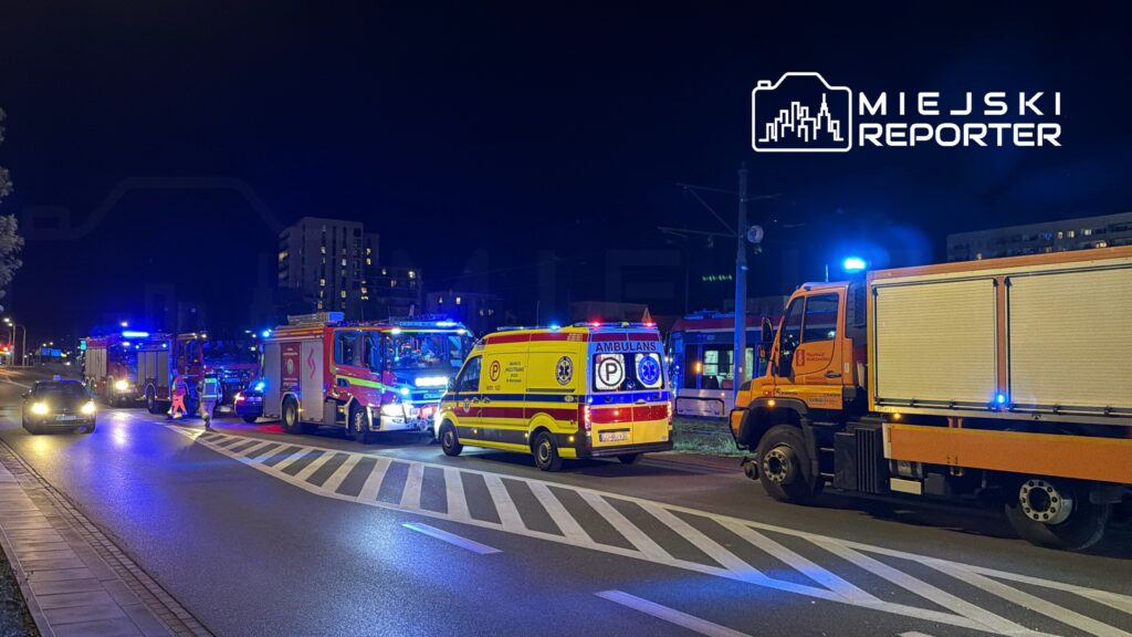 W nocy na ulicy zatrzymane są pojazdy służb ratunkowych, w tym karetka i wozów strażackich, z włączonymi sygnałami świetlnymi.