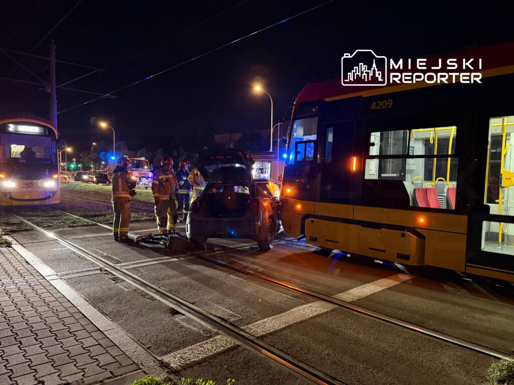 Grupa strażaków sprawdza uszkodzony samochód obok tramwaju w nocy, w tle widać pojazdy ratunkowe.
