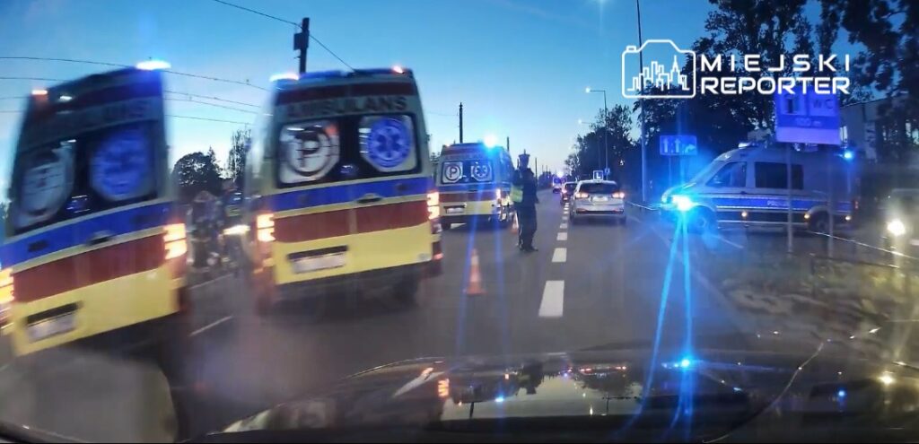 Karetki z włączonymi sygnałami świetlnymi zatrzymane na drodze, a policjant kierujący ruchem w nocnym otoczeniu.