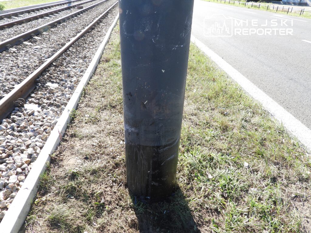 Metalowy słup przy torach kolejowych, otoczony trawą i żwirem, w tle widoczna asfaltowa droga.