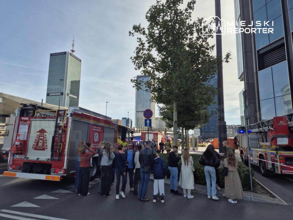 Grupa ludzi obserwuje akcję ratunkową przy wozie strażackim na ruchliwej ulicy w mieście, w tle nowoczesne wieżowce.