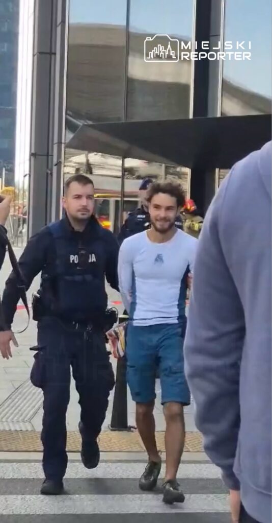 Mężczyzna w niebieskich szortach i białej koszulce jest prowadzony przez policjanta na miejskiej ulicy.