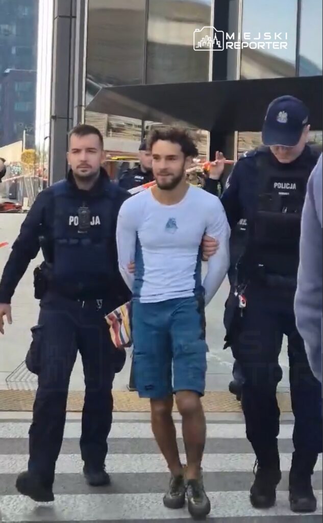 Mężczyzna w jasnej koszulce i szortach prowadzony przez dwóch policjantów na przejściu dla pieszych.