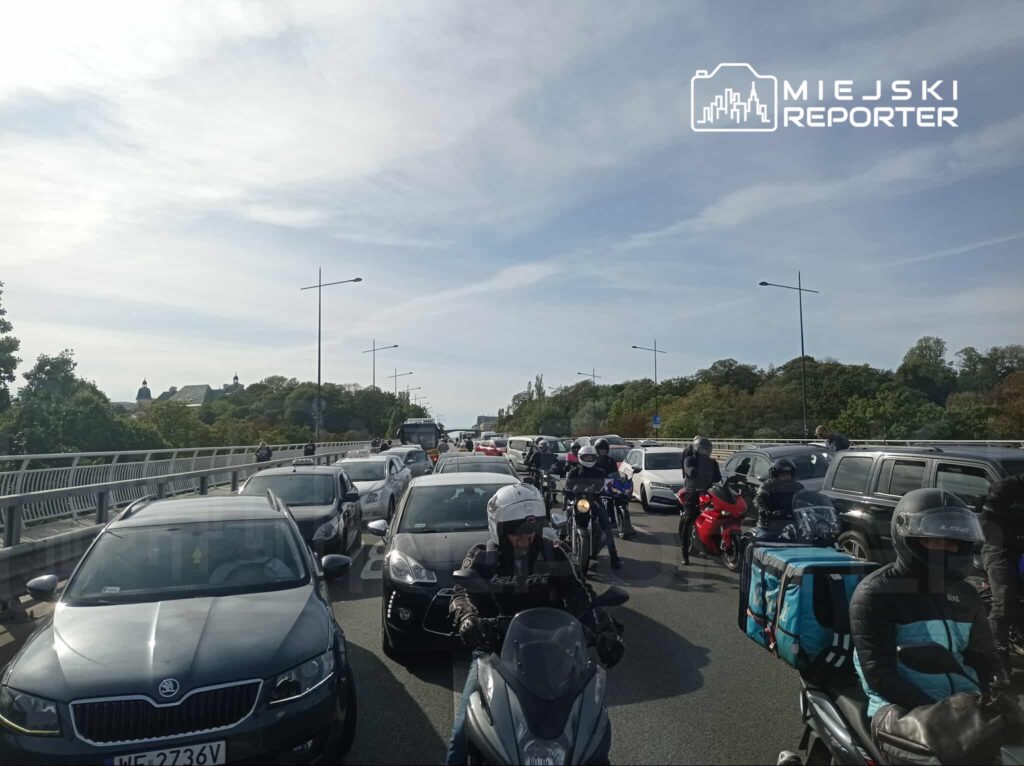 Ruch samochodowy oraz motocykle utknęły w korku na moście, w tle widać zieleń drzew i niebo.