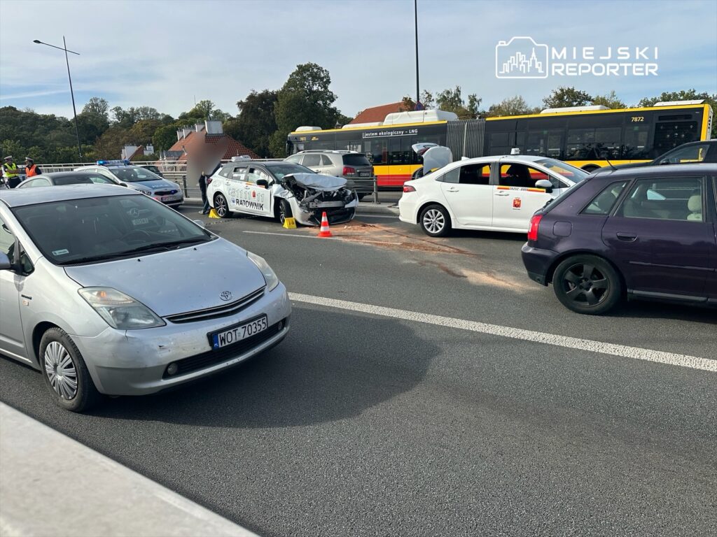 W miejscu wypadku drogowego ratownicy udzielają pomocy poszkodowanym, obok uszkodzony samochód i stojące pojazdy.