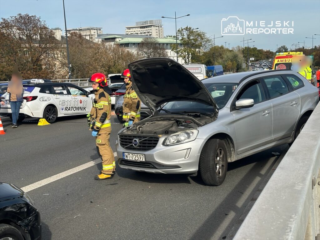 Grupa strażaków w mundurach sprawdza uszkodzony samochód osobowy na ruchliwej drodze, obok stoi taksówka.