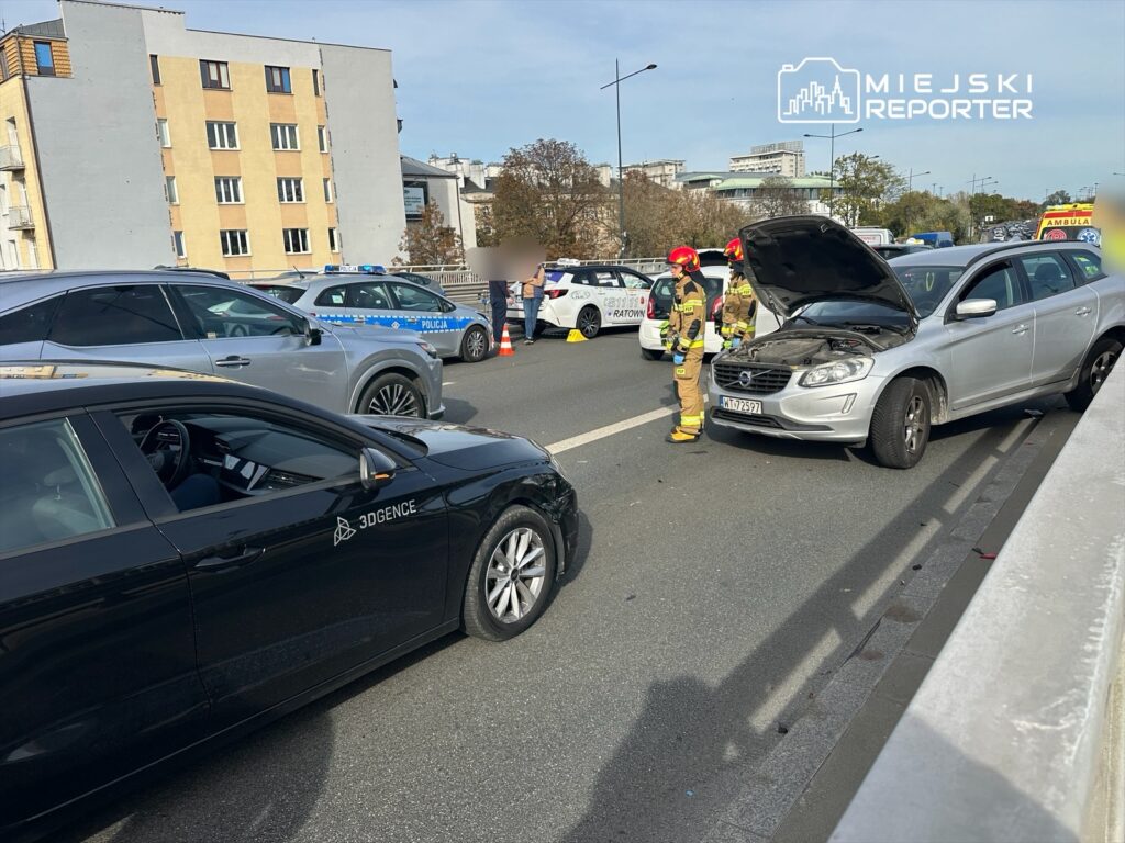 Na jezdni zatrzymane dwa samochody, w tym srebrny pojazd z otwartą maską, obok stoją strażacy w mundurach.