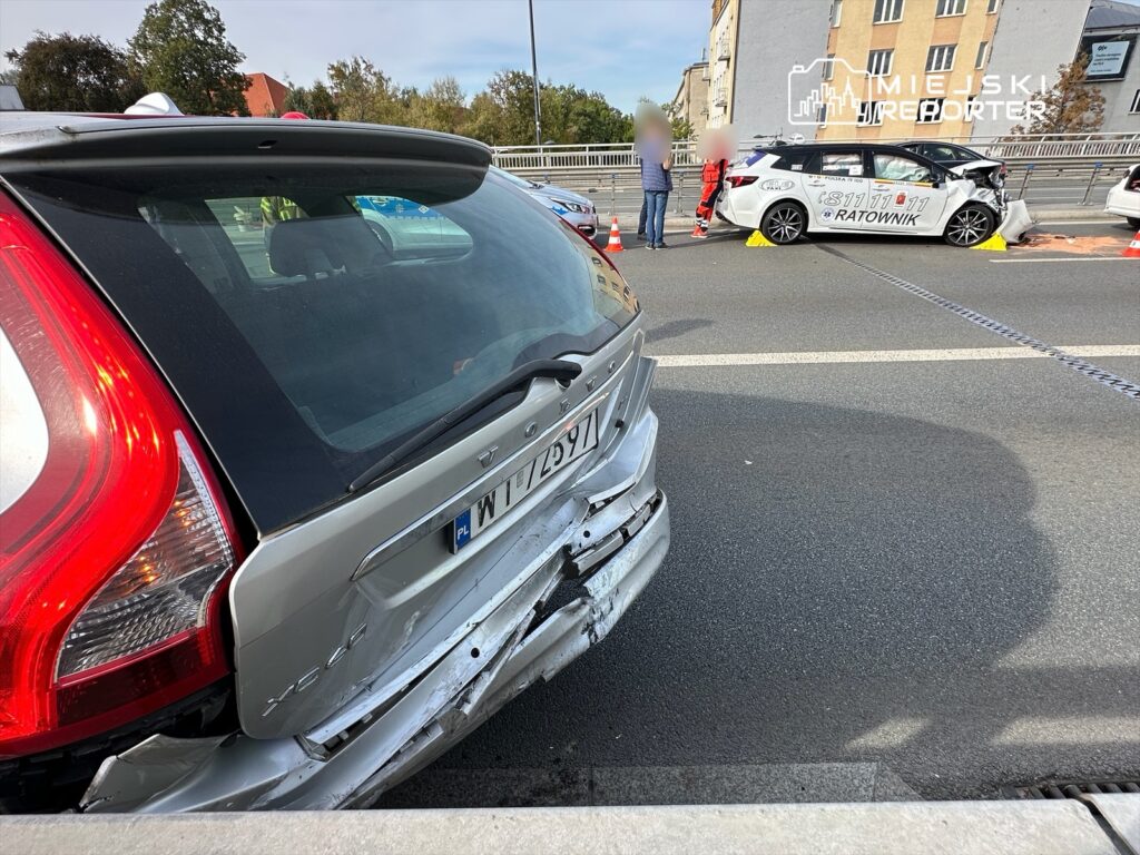 Uszkodzony samochód osobowy na drodze, obok którego znajdują się służby ratunkowe zabezpieczające miejsce zdarzenia.