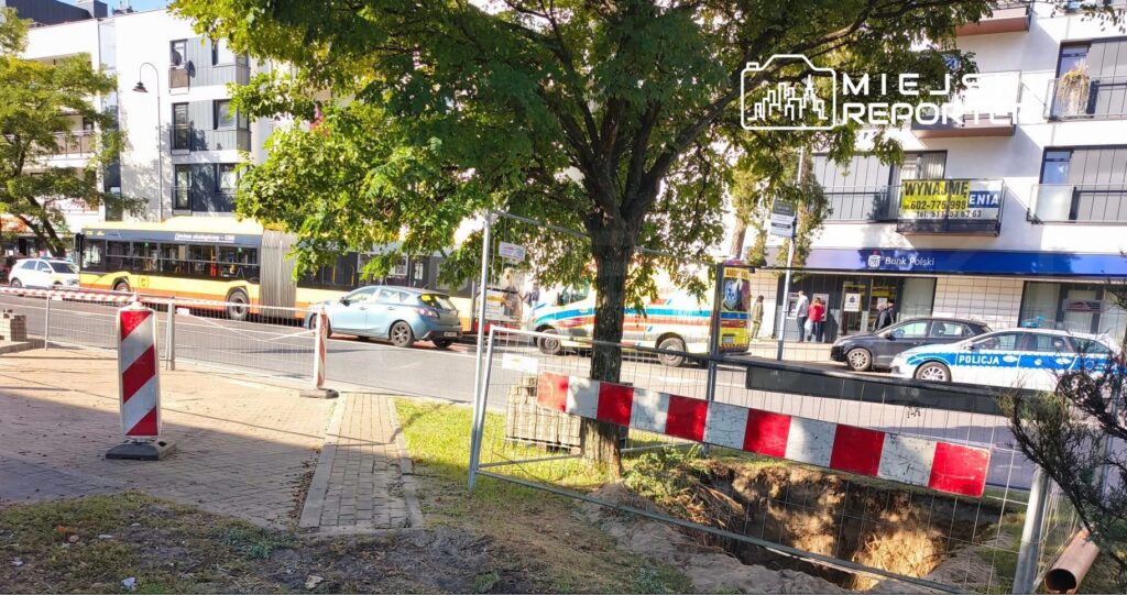 Ruch uliczny w pobliżu wykopu budowlanego, z autobusem, samochodami osobowymi i radiowozem policji na drodze.